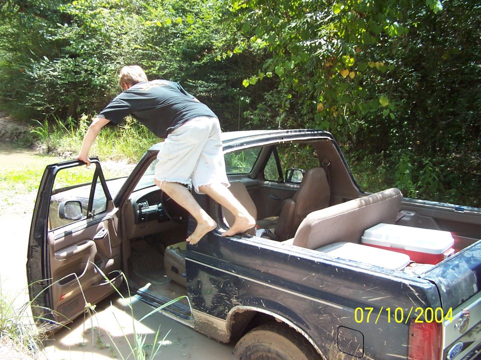 Man climbing into a muddy 1987 Ford Bronco with the driver's door open. Two coolers are in the back
