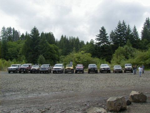 1994 Ford Explorer - Several off-road vehicles are lined up in a gravel lot, with a dense forest behind them under a cloudy sky.