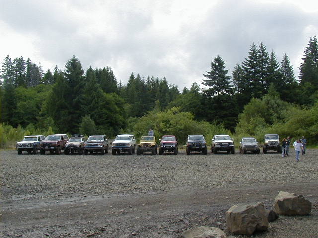 Row of lifted 4x4 trucks and SUVs parked on gravel at a forest trailhead
