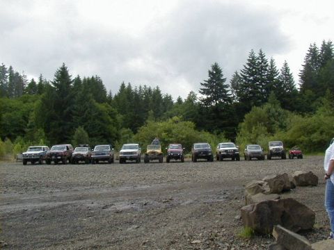1994 Ford Explorer - Multiple SUVs and trucks are parked in a line on a gravel lot, with a dense forest behind them under a cloudy sky.
