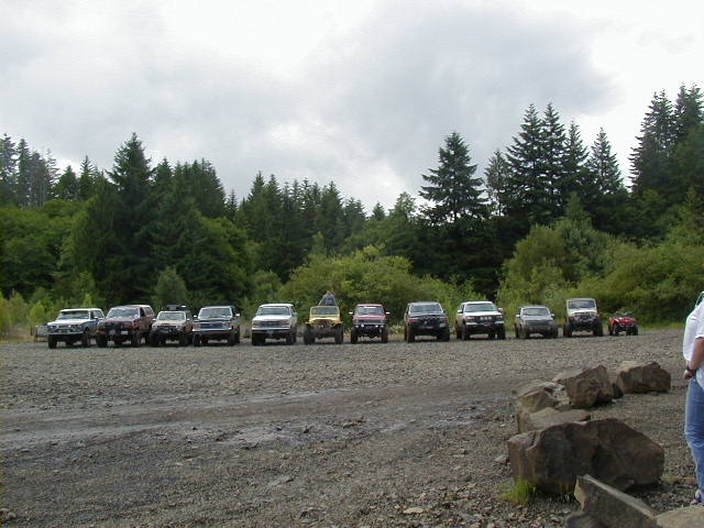 Line of SUVs and trucks parked on a gravel lot with trees in the background