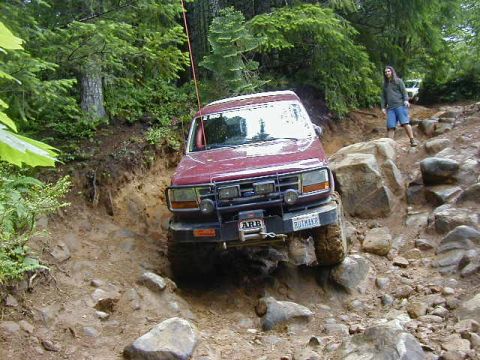 1994 Ford Explorer - A red SUV navigates a challenging rocky trail, with a person standing on the side observing.