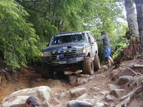 1994 Ford Explorer - A dark blue Ford Bronco navigates a challenging, rocky trail surrounded by trees.