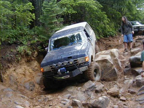 1994 Ford Explorer - A dark blue SUV navigates a challenging rocky and muddy off-road trail, with another vehicle visible in the background.