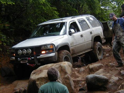1994 Ford Explorer - A white SUV tackles a challenging off-road trail with large rocks and mud, while another vehicle follows.