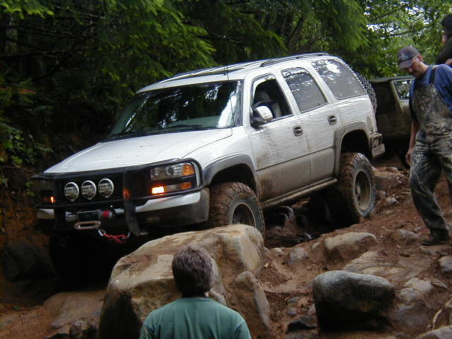 White SUV with off-road tires navigating a difficult rocky path; another vehicle is partially visible behind it