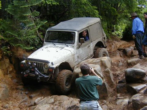 1994 Ford Explorer - A Jeep Wrangler TJ tackles a challenging rocky and muddy off-road course with spectators watching.