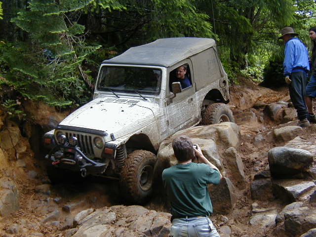 Jeep Wrangler TJ with a hardtop navigating a steep, rocky, and muddy off-road trail. A person is taking a photo of the Jeep