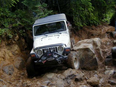 1994 Ford Explorer - A white Jeep Wrangler tackles a challenging off-road course, with large rocks and mud obscuring the path.