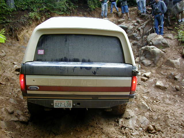 Rear view of a tan and grey Ford pickup truck with a camper shell, driving on a rocky, muddy trail