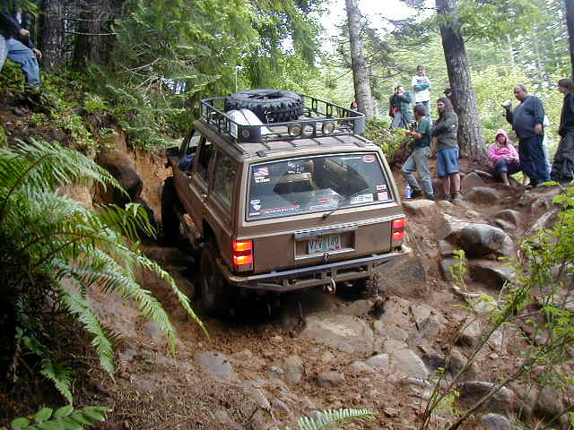 Brown Jeep Cherokee XJ driving up a rocky, wooded trail with people watching