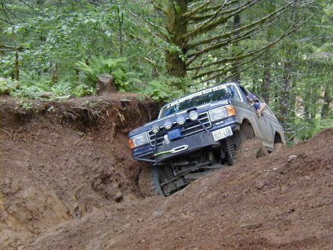 1994 Ford Explorer - An off-road SUV navigates a challenging dirt incline, its tires digging into the loose soil amidst a dense forest.