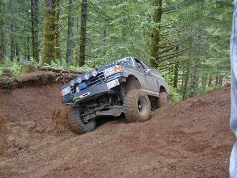 1994 Ford Explorer - A modified Ford Bronco navigates a steep, muddy incline in a wooded area.