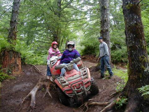 1994 Ford Explorer - Two people ride an ATV through a wooded area with muddy terrain and large trees.