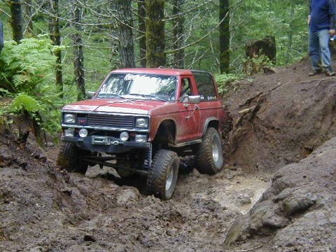 1994 Ford Explorer - A red Ford Bronco navigates a challenging muddy trail in a wooded area.