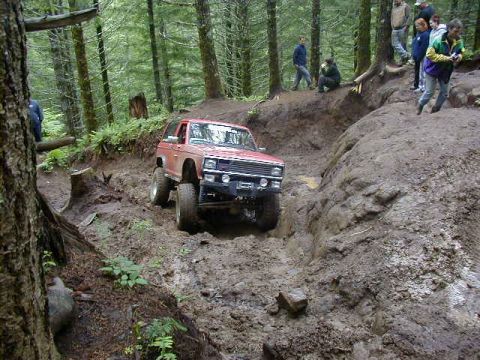 1994 Ford Explorer - A red SUV tackles a challenging muddy trail during an off-road event, with spectators watching from the embankment.