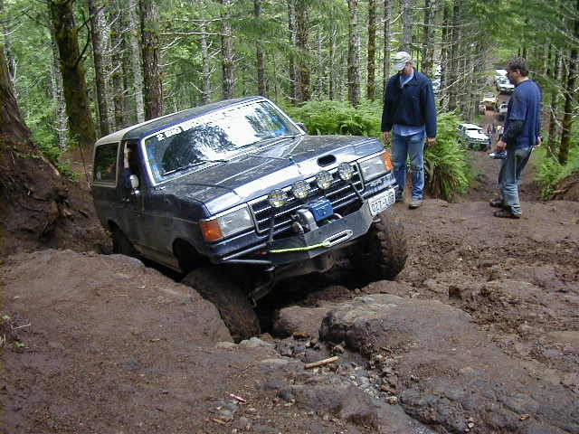 Dark blue off-road SUV with aftermarket bumper and lights stuck on a dirt trail, two people watching