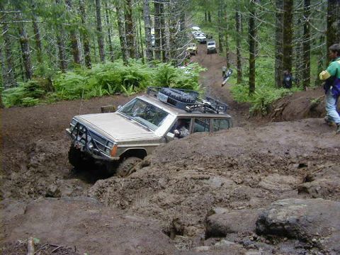 1994 Ford Explorer - A tan SUV navigates a challenging muddy obstacle on a wooded trail, with several other vehicles following.