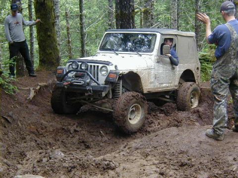 1994 Ford Explorer - A Jeep Wrangler tackles a challenging muddy off-road course with spectators watching.