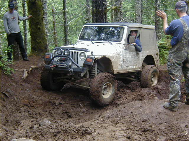 White Jeep Wrangler with soft top navigating a muddy trail, two men nearby
