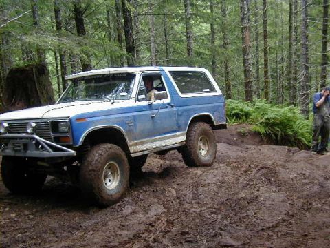 1994 Ford Explorer - A blue and white Ford Bronco navigates a muddy off-road trail surrounded by dense forest.