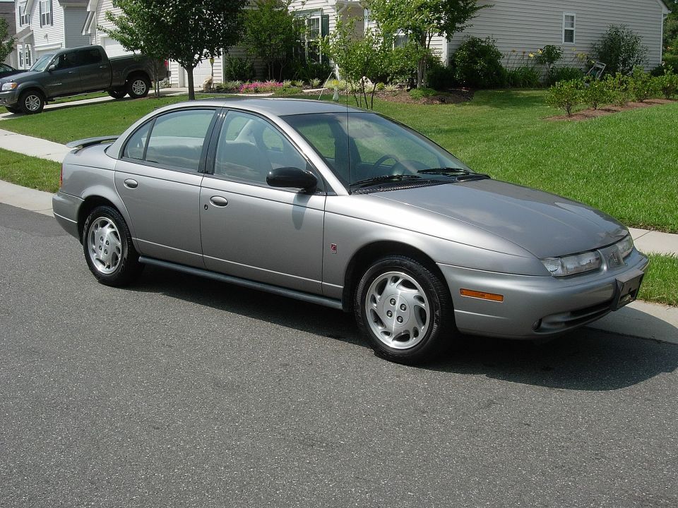 Silver Saturn sedan parked on street, with a pickup truck on a lawn in the background