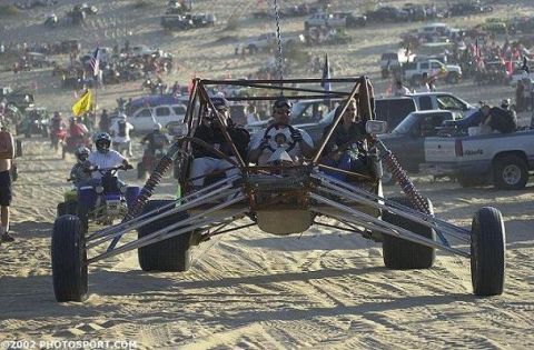 1990 Ford Bronco - three-seat tube-frame sand rail crests a sandy dune surrounded by spectators, ATVs and parked trucks.