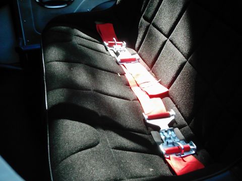 1978 Ford Bronco - The interior of a vehicle shows a black racing seat with a red five-point harness system.