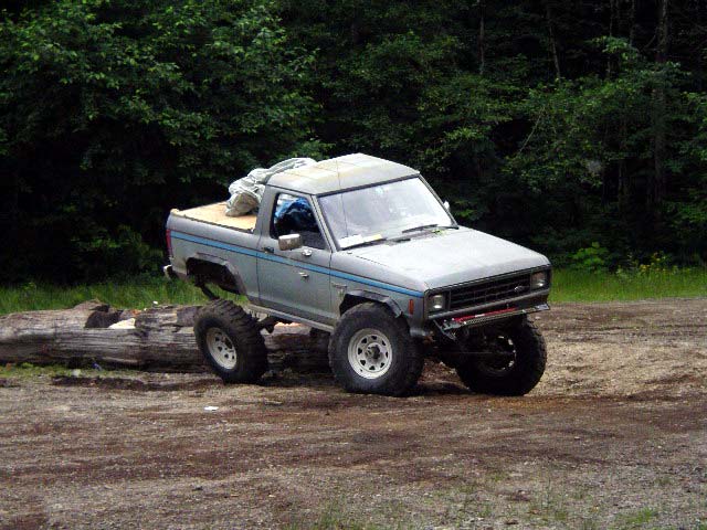 Gray Ford Ranger pickup truck with lifted suspension and off-road tires, parked on dirt next to a log
