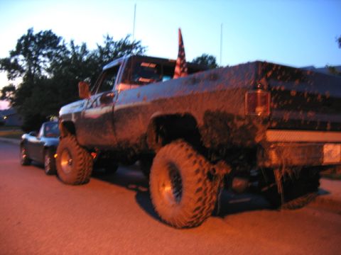 1976 GMC C/K Pickup - A heavily muddied pickup truck is parked on a street, with a smaller car behind it.