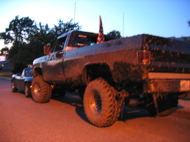 Mud-covered lifted square-body pickup with American flag parked at dusk, small roadster ahead
