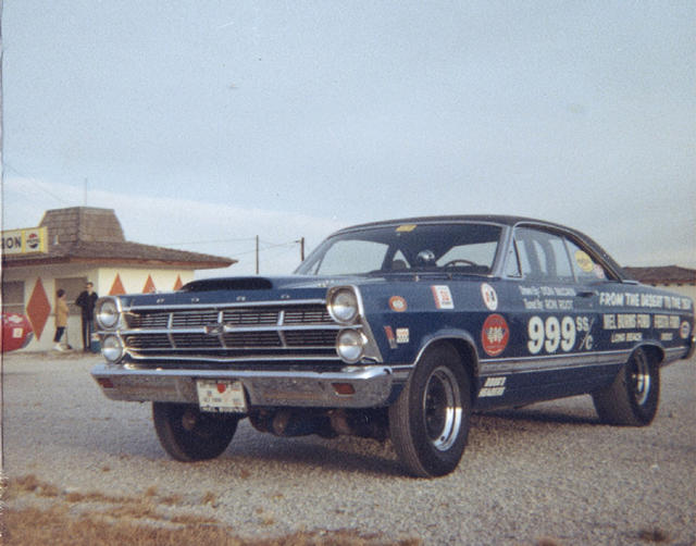 Vintage blue drag-racing coupe with race lettering parked on gravel