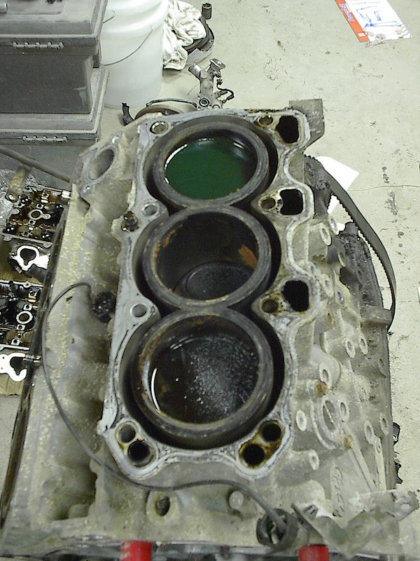 Disassembled engine block with three cylinder bores, one filled with green fluid, on a shop floor