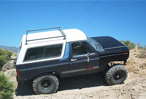 1986 Ford Bronco - This Ford Bronco is equipped with off-road tires and a roof rack, navigating a sandy terrain under a clear blue sky.