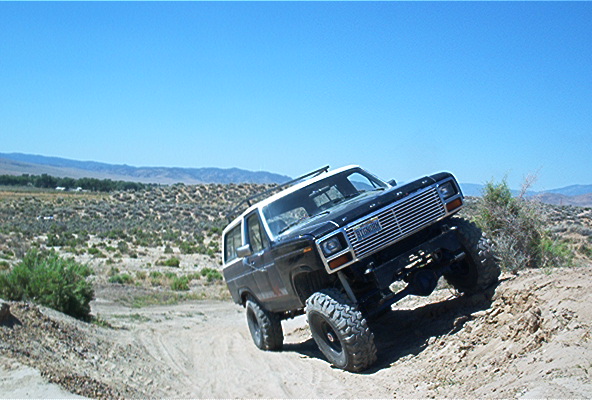 Black Ford Bronco with lifted suspension driving on a dirt hill. Chrome grille and white roof are visible