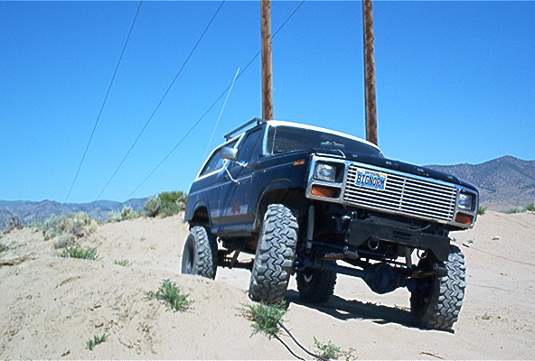 1986 Ford Bronco with large tires climbing a sandy hill under a clear blue sky