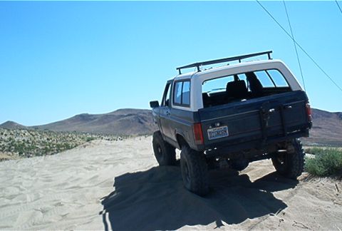1986 Ford Bronco - This 1986 Ford Bronco is captured from the rear as it navigates a sandy terrain under a clear blue sky.