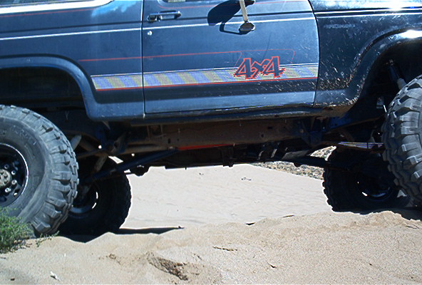 Blue Ford Bronco with 4x4 decal, lifted suspension, and large tires on sandy terrain