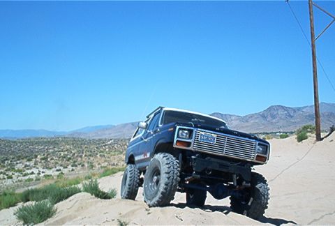 1986 Ford Bronco - This lifted 1986 Ford Bronco navigates a sandy, off-road landscape under a clear blue sky.