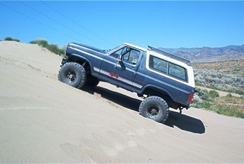 1986 Ford Bronco - A blue and white Ford Bronco navigates a sandy incline under a clear blue sky.