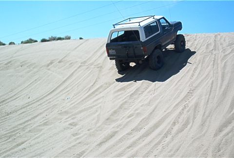 1986 Ford Bronco - This lifted Ford Bronco is navigating a steep sand dune under a clear blue sky.