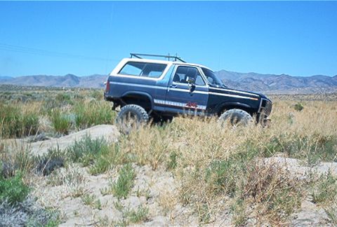 1986 Ford Bronco - This 1986 Ford Bronco navigates a sandy incline in a desert landscape under a clear blue sky.