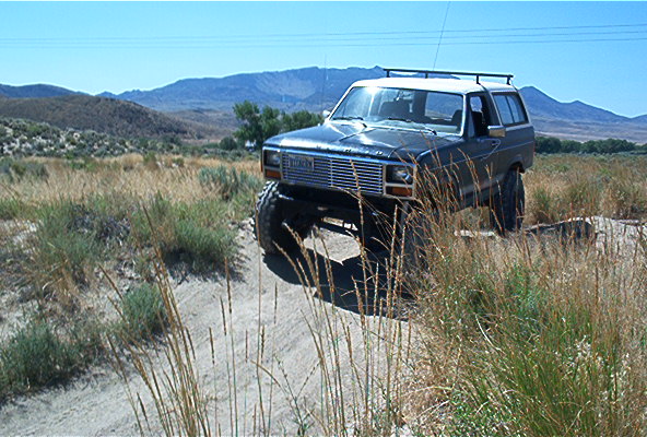1986 Ford Bronco with lifted suspension driving on a dirt trail with mountains in the background