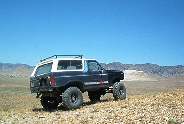 Dark blue and white Ford Bronco with off-road tires parked on a dry, grassy hill under a clear blue sky