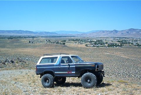1986 Ford Bronco - A lifted blue and white SUV with a roof rack sits in a dry, hilly landscape under a clear blue sky.