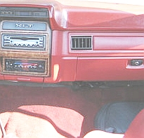 1986 Ford Bronco - Interior view of a red dashboard and glovebox area with woodgrain trim and an aftermarket head unit mounted in the center stack.