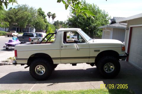 1995 Ford Bronco - Driver-side view of a cream/white lifted Ford full-size Bronco with the hardtop removed and a black interior roll bar, parked in a resident…
