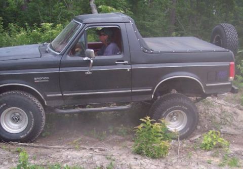 1988 Ford Bronco - Black full-size Ford Bronco with the hardtop removed and a black tonneau cover over the rear, driven by a man in a ball cap on a dirt trail…