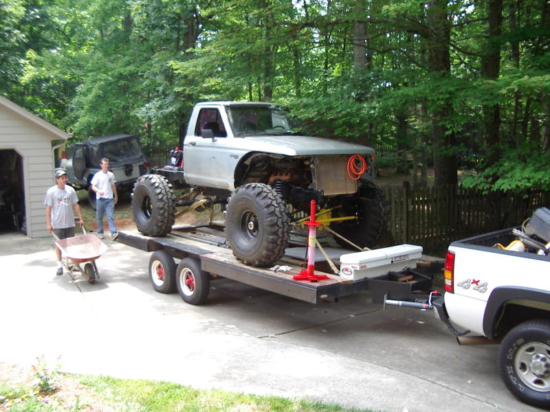 Silver pickup truck chassis with large tires on a trailer, being loaded by two men. Another SUV is parked nearby