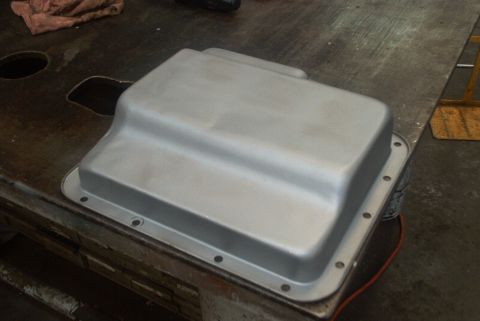 1983 Ford Bronco - A silver transmission oil pan sits on a dark, stained workbench, with a red wire peeking out from beneath it.